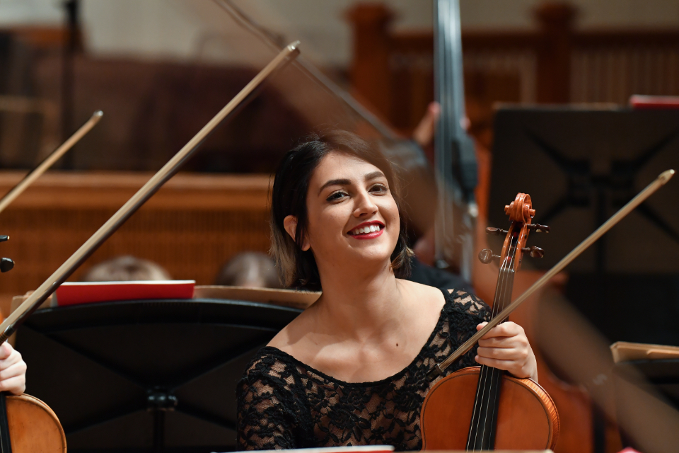 A female student, with short dark hair, smiling, while holding her violin, sitting in an orchestra with other violins.